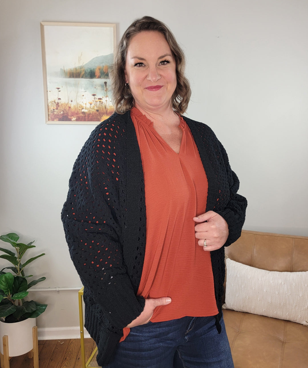 A woman with wavy brown hair smiles indoors, wearing the ee:some Margot Open Front, Open Knit Cardigan over a rust blouse and jeans. Behind her are a framed painting, green plant, tan sofa, and white pillow. Perfect for stylish layering.