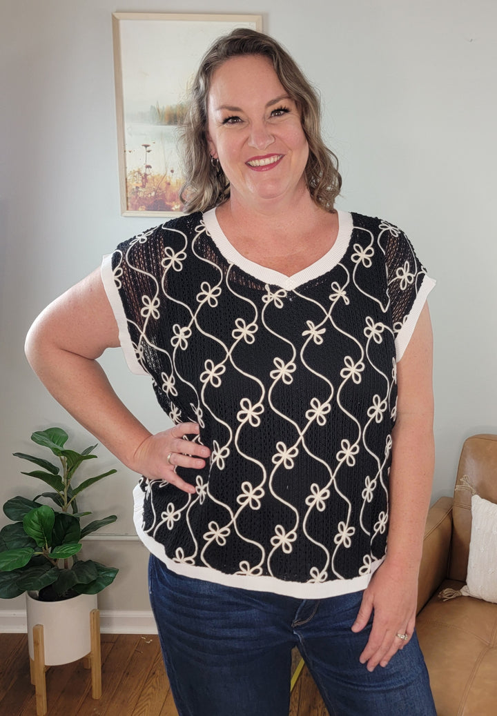 A smiling woman with wavy brown hair poses indoors wearing the ODDI Zoie Floral Embroidered V-Neck Top. She stands with one hand on her hip, a plant and framed artwork visible in the background.
