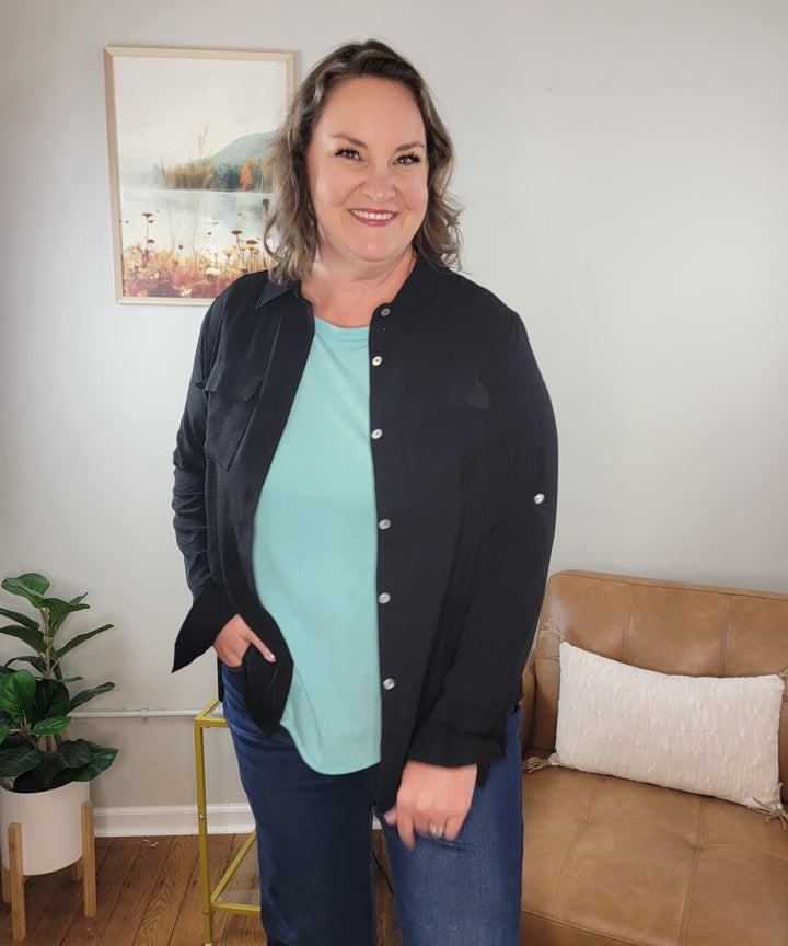 A smiling woman with wavy brown hair stands indoors near a tan couch and potted plant, wearing the Spin James Button Down Top in black over a light blue lightweight top and jeans. A framed nature picture hangs on the wall behind her.