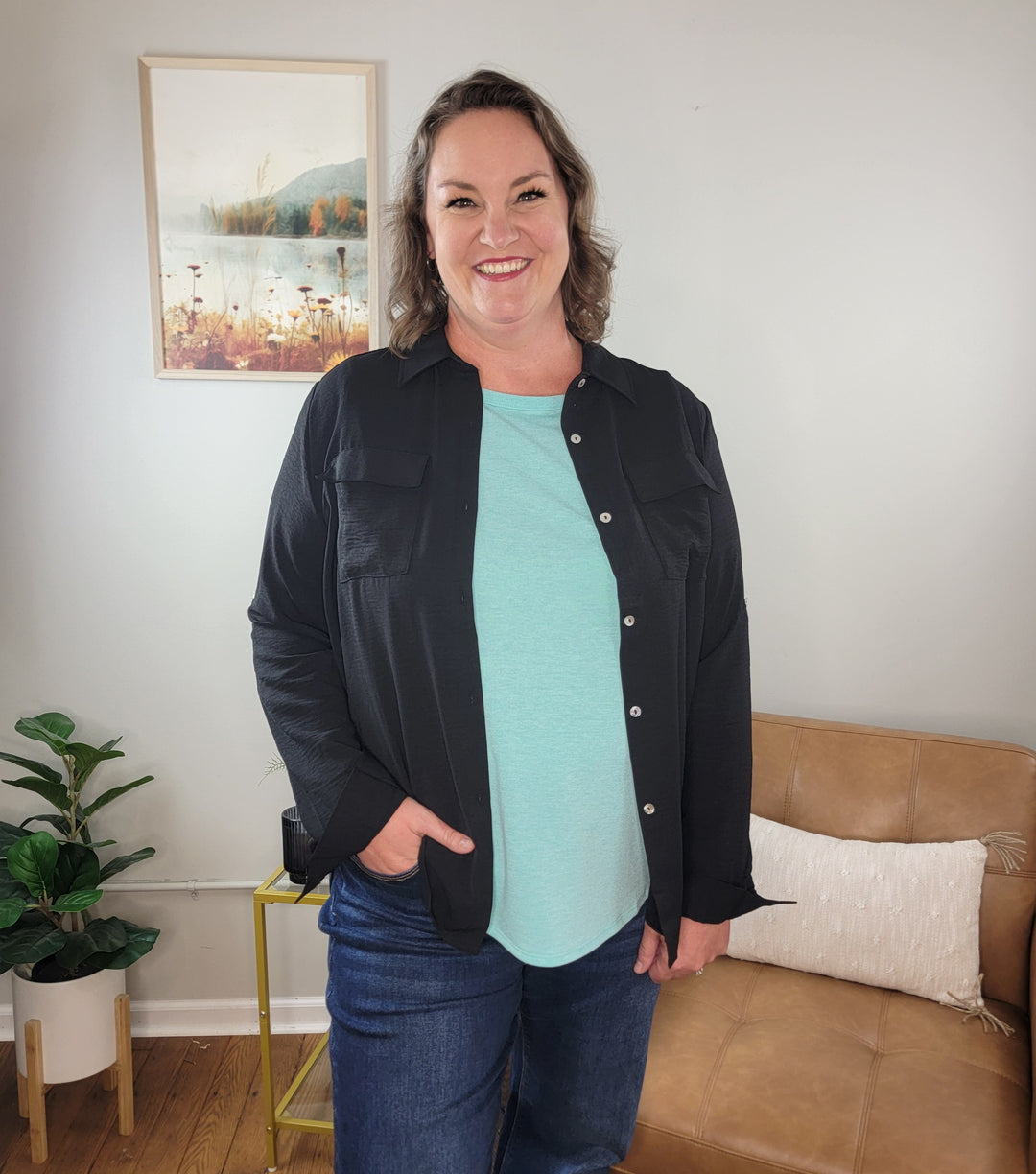 A woman smiles indoors by a tan couch with a pillow, wearing the Spin James Button Down Top over a teal shirt and blue jeans. Behind her are a framed nature photo and a potted plant on the wooden floor.