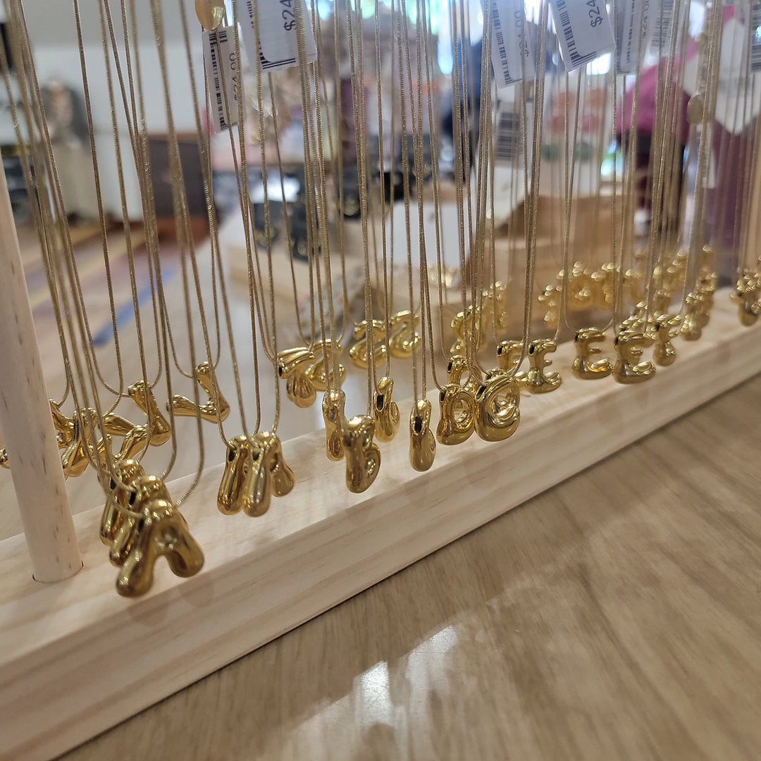 Cape Diablo Bubble Letter Necklaces hang on thin chains from a wooden display stand, arranged neatly in rows. The gold pendants form alphabet letters, with a blurred retail store background.