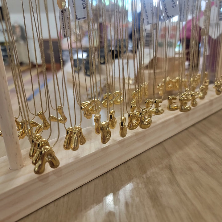 Cape Diablo Bubble Letter Necklaces hang on thin chains from a wooden display stand, arranged neatly in rows. The gold pendants form alphabet letters, with a blurred retail store background.