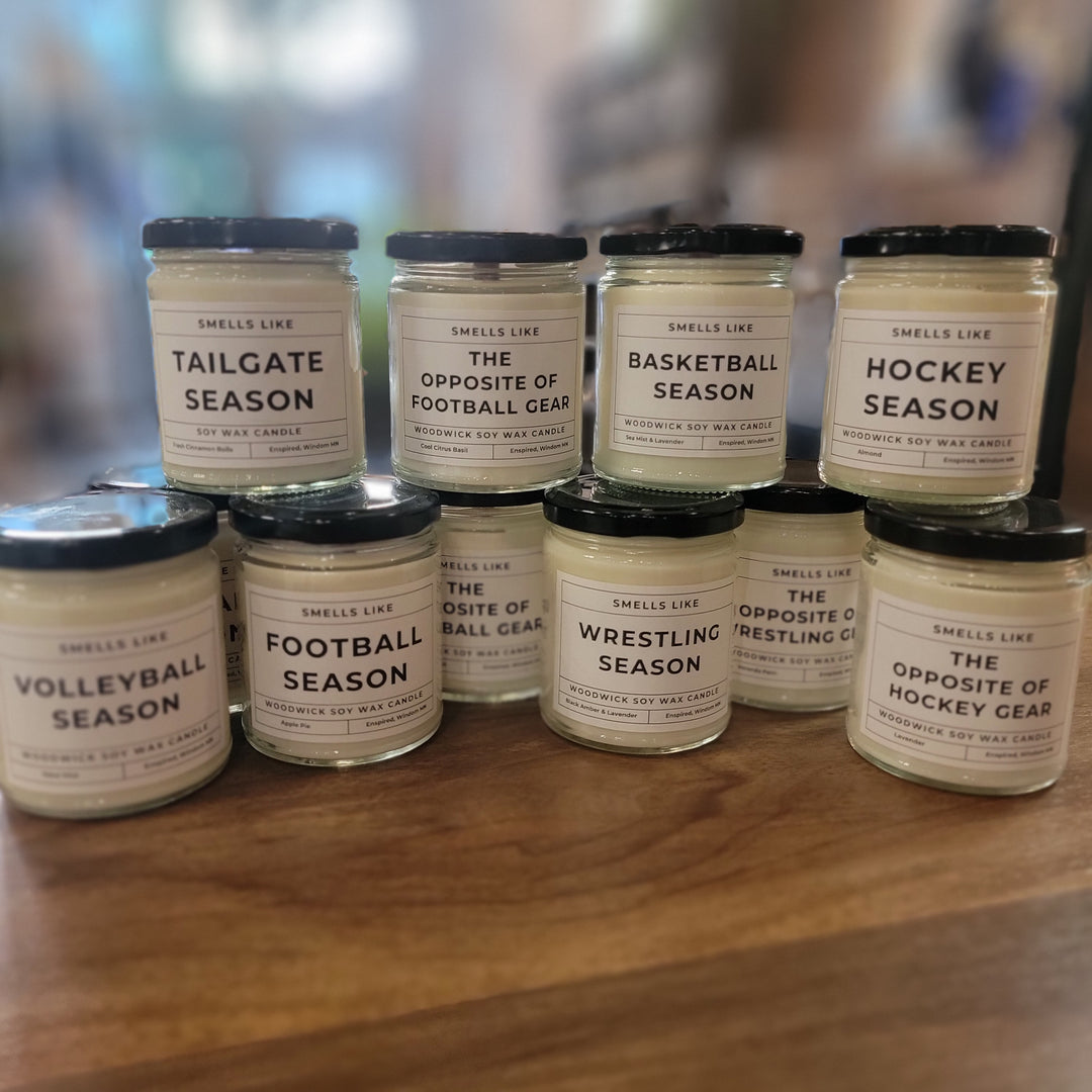 Nine Cosmos + Clover Boutique Sports Candles are displayed on a wooden surface. Each soy wax candle features custom labels for different sports seasons, including tailgate, basketball, football, hockey, volleyball, and wrestling. A blurred background is visible.