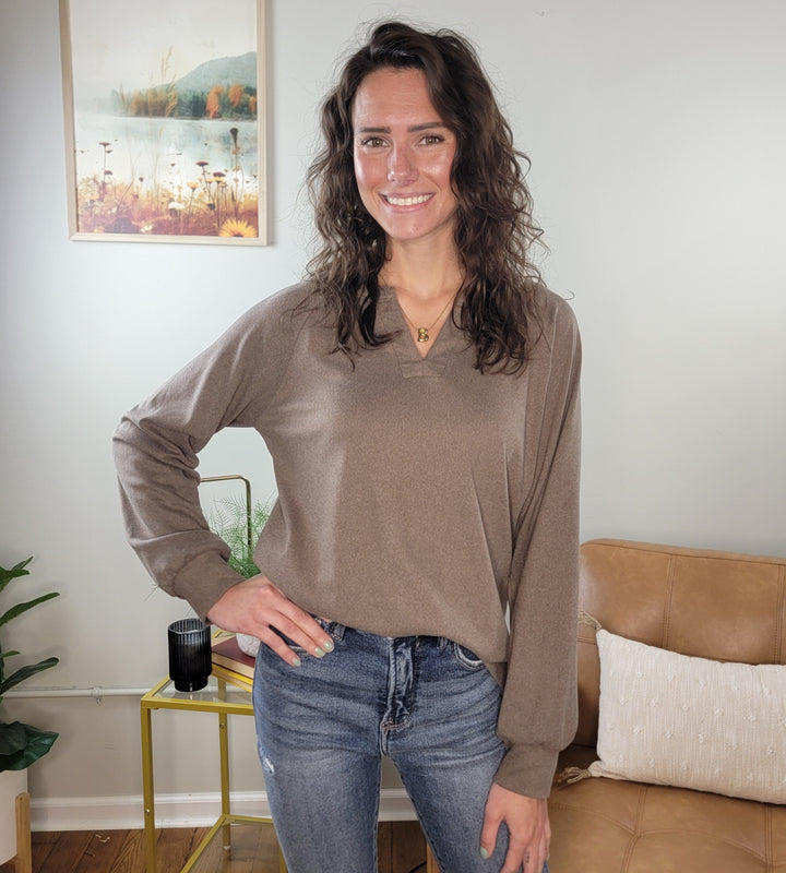 A woman with wavy brown hair smiles indoors by a tan couch, wearing the Staccato Eira Casual Long Sleeve V-Neck Top and jeans. She poses with one hand on her hip; a framed nature picture hangs on the wall behind her.