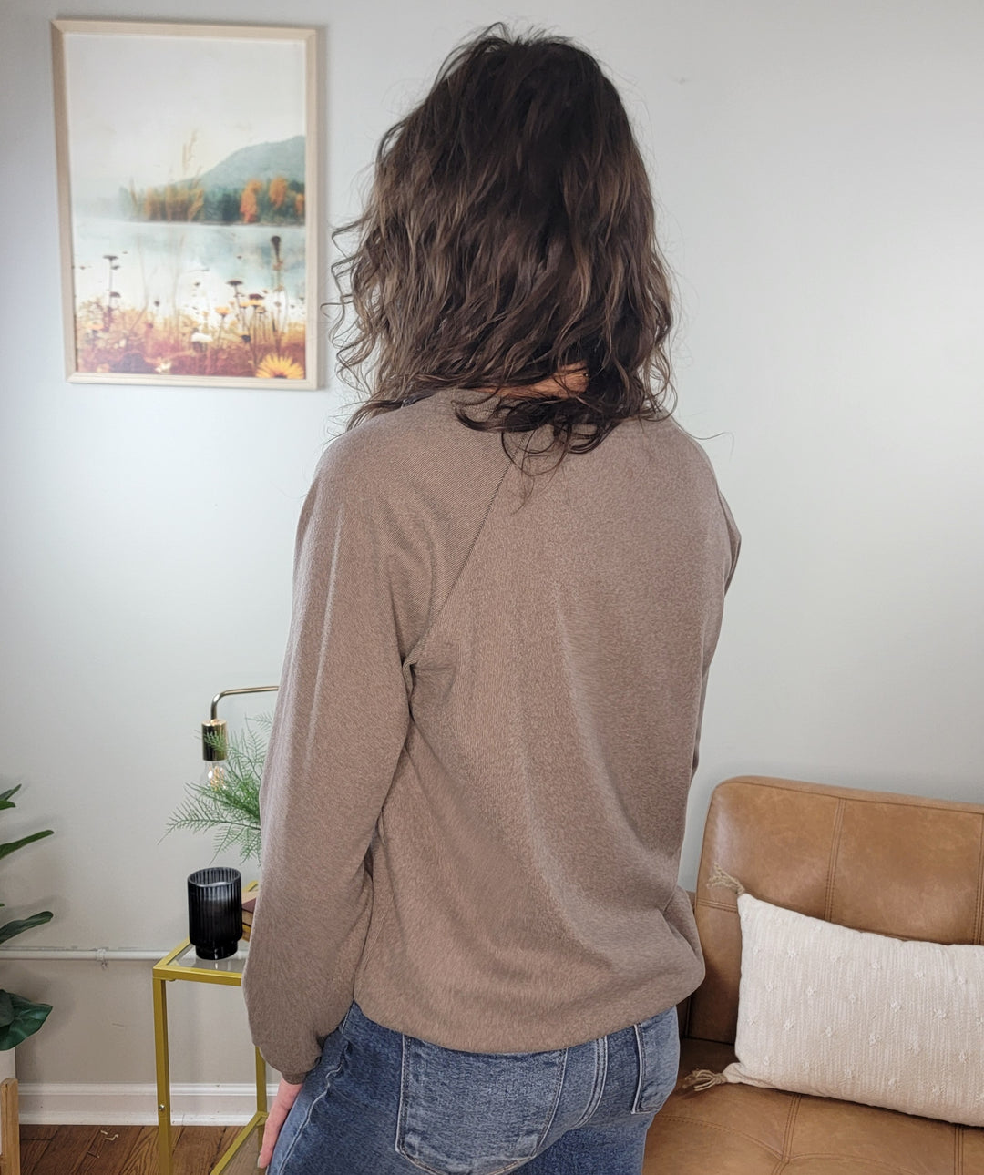 A person with wavy brown hair, wearing the Staccato Eira Casual Long Sleeve V-Neck Top and blue jeans, stands indoors facing away. The background features a beige couch, yellow side table, plant, and landscape picture on the wall.