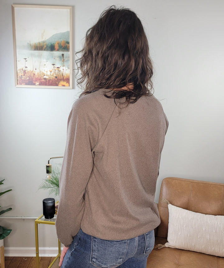 A person with wavy brown hair, wearing the Staccato Eira Casual Long Sleeve V-Neck Top and blue jeans, stands indoors facing away. The background features a beige couch, yellow side table, plant, and landscape picture on the wall.