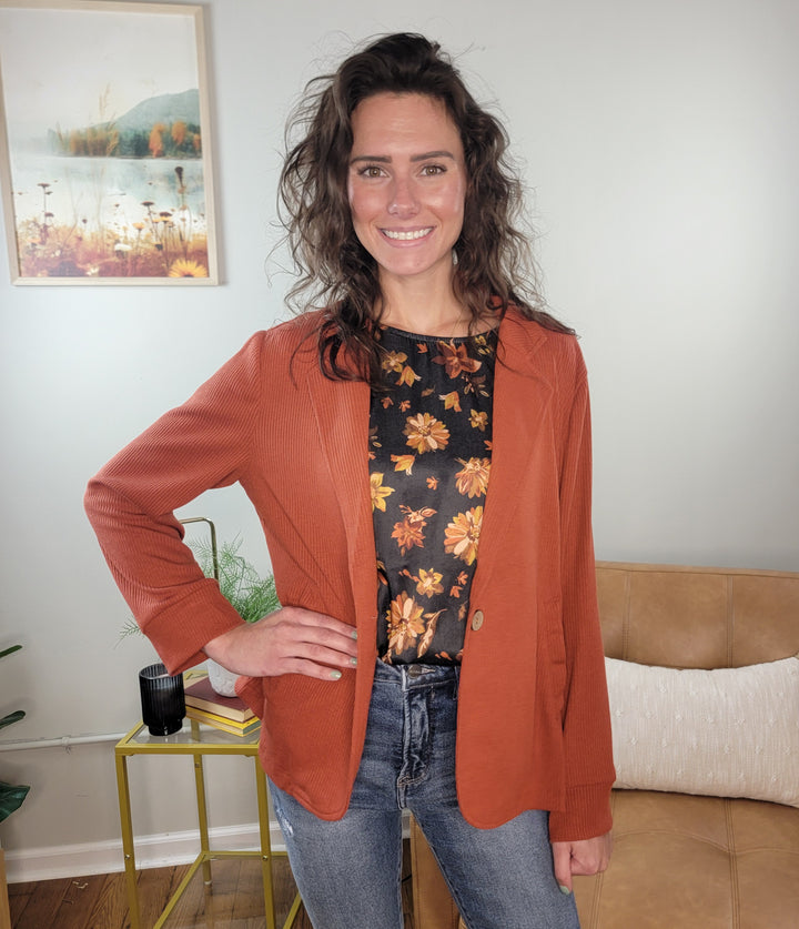 A woman with wavy brown hair smiles, wearing the Doe &amp; Rae Arlet Stretch Blazer in burnt orange over a floral black shirt and blue jeans. She stands in a modern living room, highlighting this chic professional look.