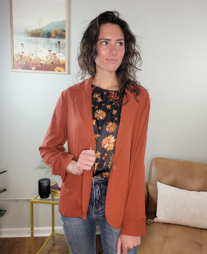 A woman with wavy brown hair stands indoors, looking to the side, wearing the Doe &amp; Rae Arlet Stretch Blazer in orange over a black floral top and jeans. Behind her are a framed floral picture and a tan chair with a pillow.