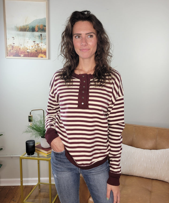 A woman with wavy brown hair stands indoors in front of a beige couch and a framed nature painting, wearing the Staccato Lucia Light Weight Striped Pullover and blue jeans.