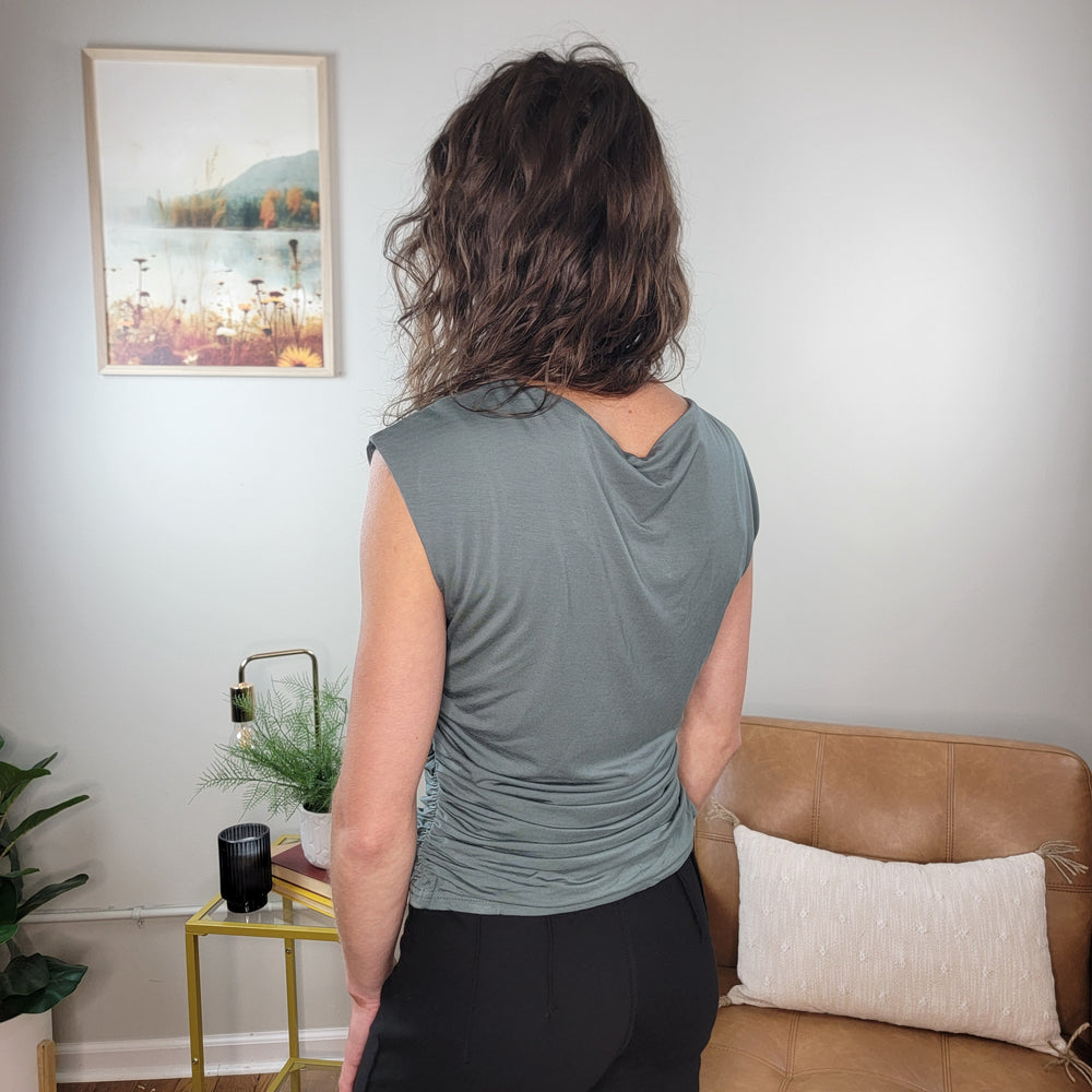 A person with wavy brown hair stands with their back to the camera, wearing the Allie Rose Piper Sleeveless Top in gray and black pants in a modern living room featuring a tan couch, yellow side table, and framed wall art.