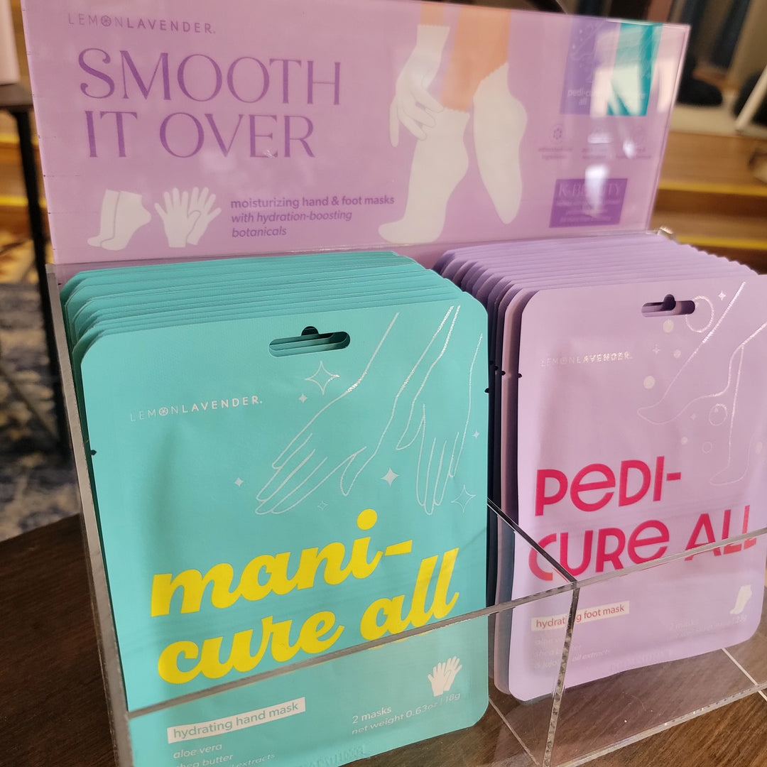 A display of DM Merchandising's Smooth It Over Masks, featuring lemon lavender hydrating hand and foot masks in teal and lavender manicure and pedicure packaging, with a "Smooth It Over" sign behind them.