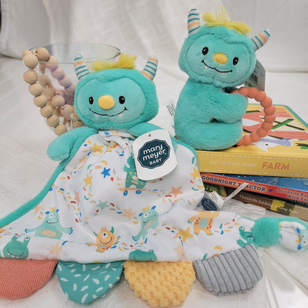 Two Mookie Monster Teether Rattle toys by Mary Meyer—a teal and yellow plush with teether ring and a blanket version—are shown beside stacked children's books and a beaded cup on a white surface.
