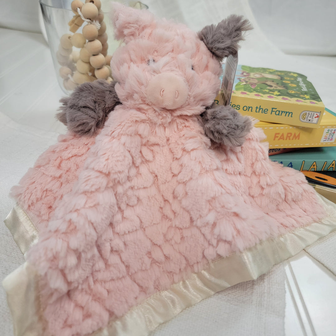 The Mary Meyer Putty Nursery Piglet Character Blanket, soft and pink with gray ears and a satin edge, is shown on a white surface with wooden beads in a glass and colorful children's books in the background.