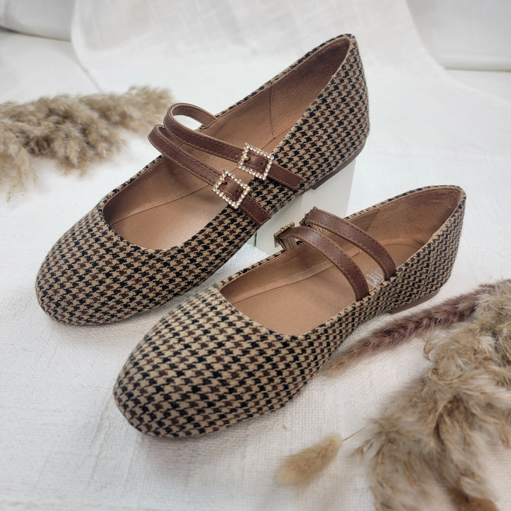 The Own It Flats from Corkys feature brown and beige houndstooth, double straps, and silver buckles, shown on white fabric with dried grasses nearby.