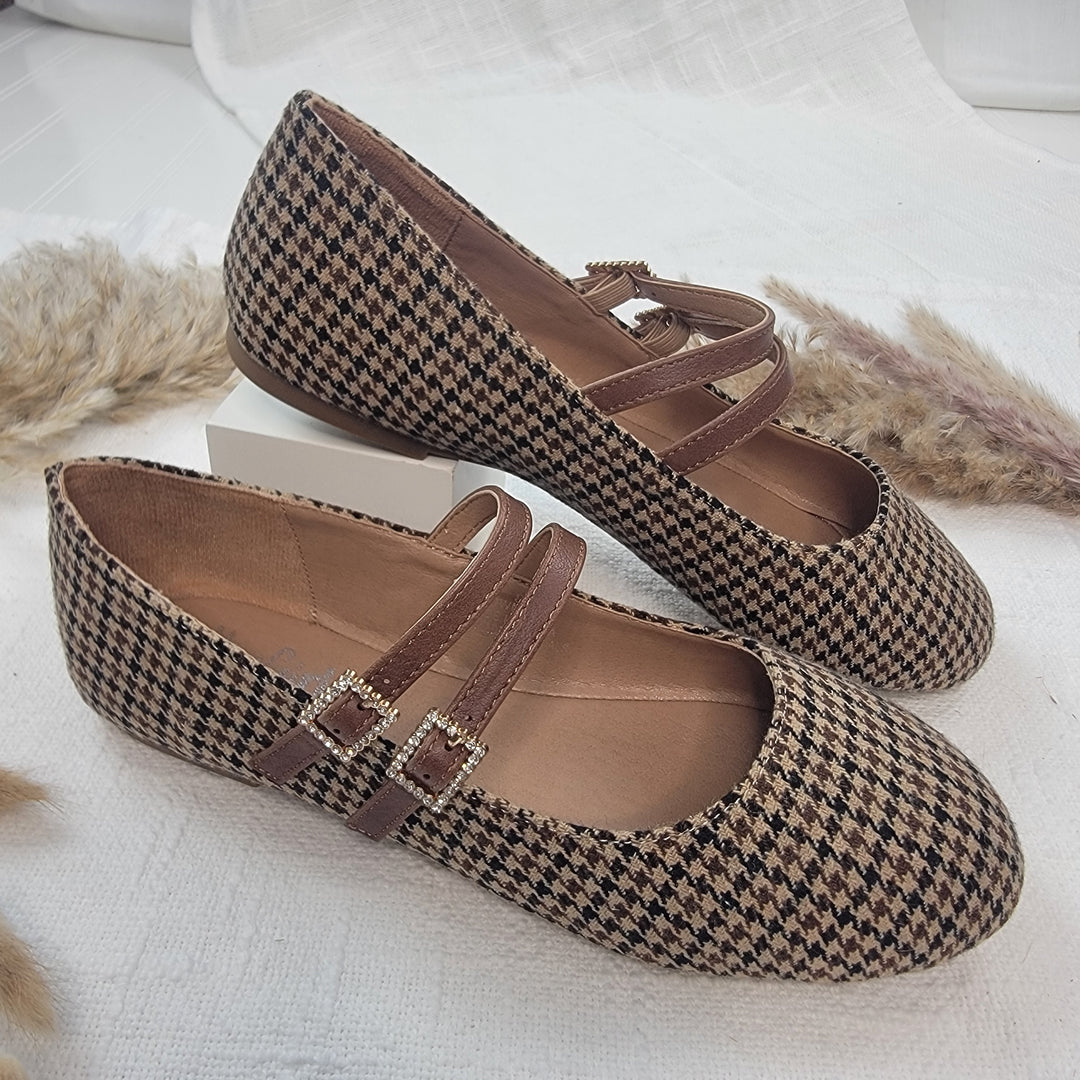 The Own It Flats from Corkys feature a flat, round-toe houndstooth design with double brown straps and rhinestone buckles, beautifully displayed on white fabric with dried grass accents.