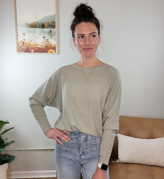A woman with curly hair in a bun stands in a living room, wearing the Allie Rose Elsie Knit Sweater in light gray and light blue jeans. She accessorizes with a necklace and smartwatch, one hand on her hip. A framed landscape hangs behind her.