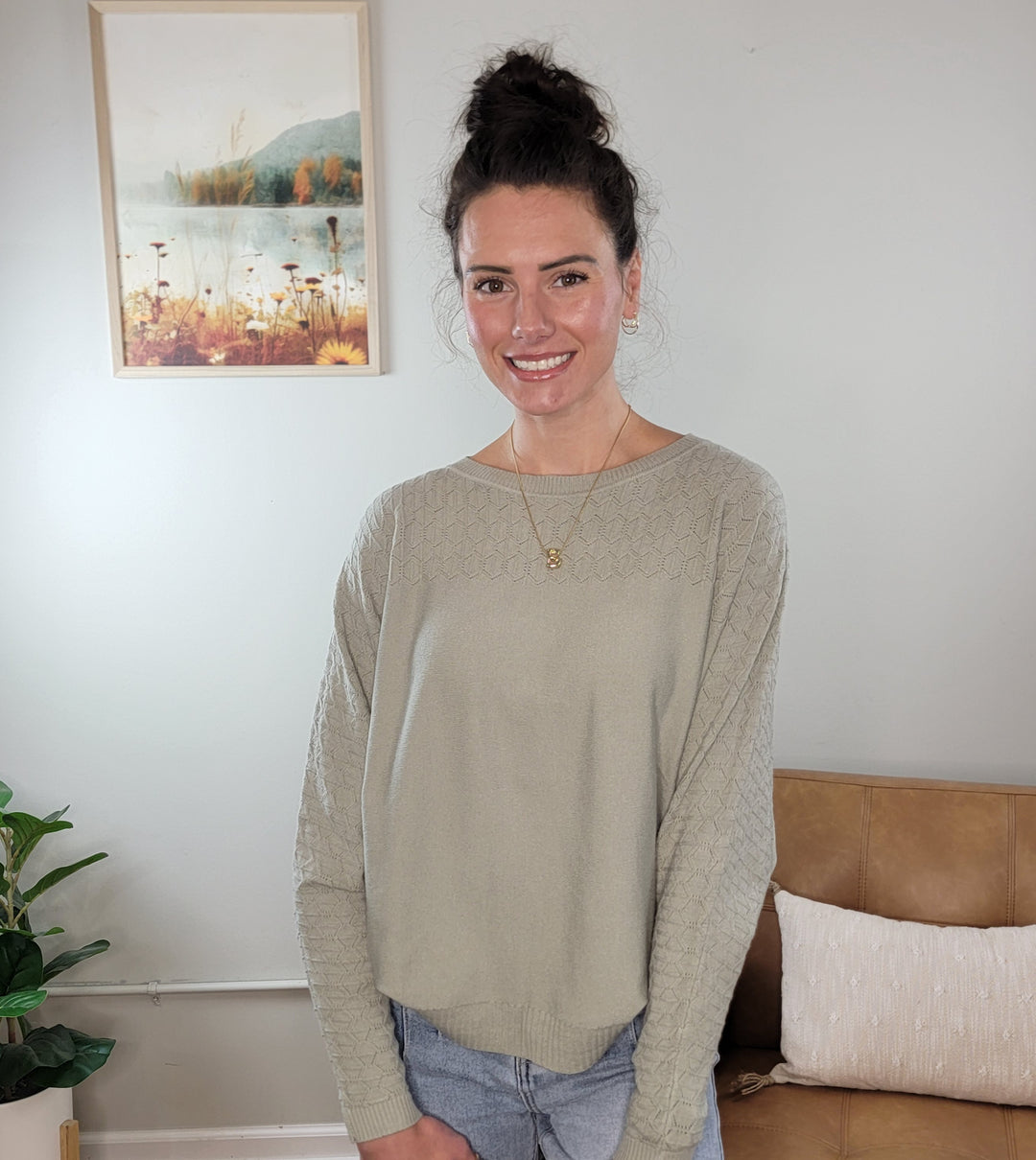 A woman with dark hair in a bun smiles at the camera indoors, wearing the Allie Rose Elsie Light Weight Spring Knit Sweater in sage green and jeans. She stands before a beige wall with a nature painting, plant, and couch visible in the background.