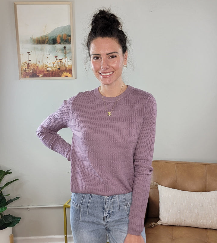 A woman with dark hair in a bun smiles indoors wearing the Allie Rose Annie Light Weight Spring Knit Sweater in lavender and light blue jeans, standing by a beige couch, cushion, leafy plant, and framed nature photo on a light gray wall.