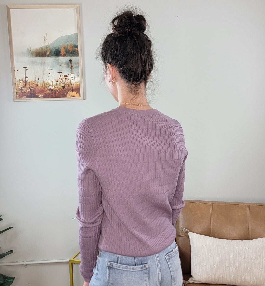 A woman with dark hair in a messy bun, wearing the Allie Rose Annie Light Weight Spring Knit Sweater in lavender and light blue jeans, stands facing away from the camera in a room with a beige couch, cushion, and landscape painting.