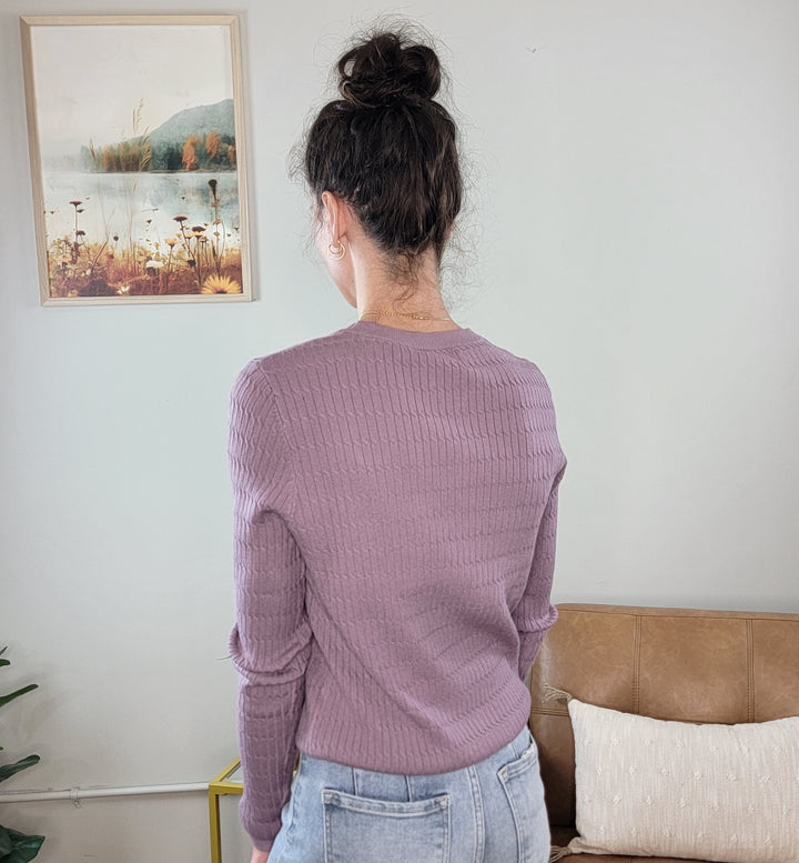 A woman with dark hair in a messy bun, wearing the Allie Rose Annie Light Weight Spring Knit Sweater in lavender and light blue jeans, stands facing away from the camera in a room with a beige couch, cushion, and landscape painting.