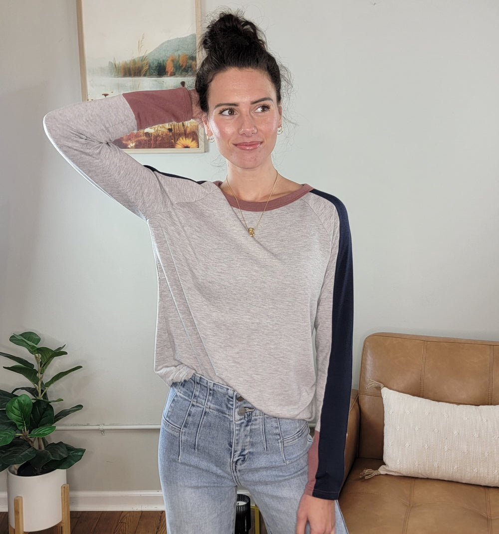 A woman with dark hair in a bun wears the Staccato Jodi Long Sleeve Top and high-waisted jeans, standing indoors with one arm resting on her head, smiling. A plant and a tan couch are visible in the background.