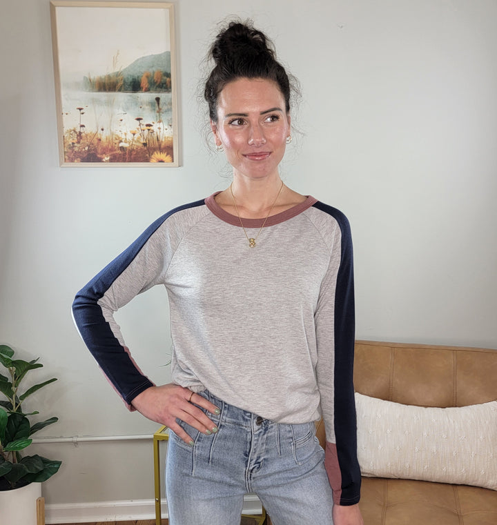 A woman with dark hair in a bun wears the Staccato Jodi Long Sleeve Top and light blue jeans, standing in a living room with a tan couch, plants, and a framed nature picture on the wall.