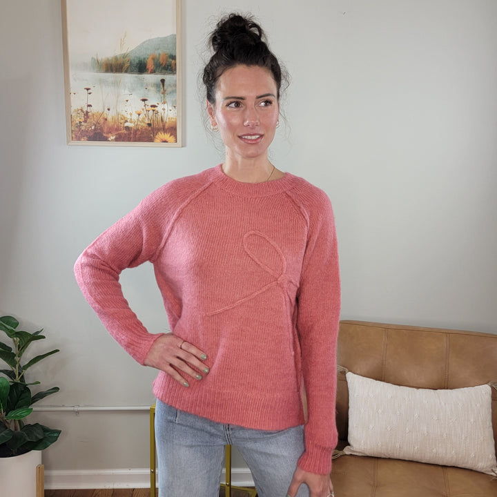 A woman, hair in a bun and smiling slightly, stands indoors with one hand on her hip wearing the Staccato Avyana Bow Embellished Knit Sweater in berry and light blue jeans. A plant and a framed picture decorate the background.