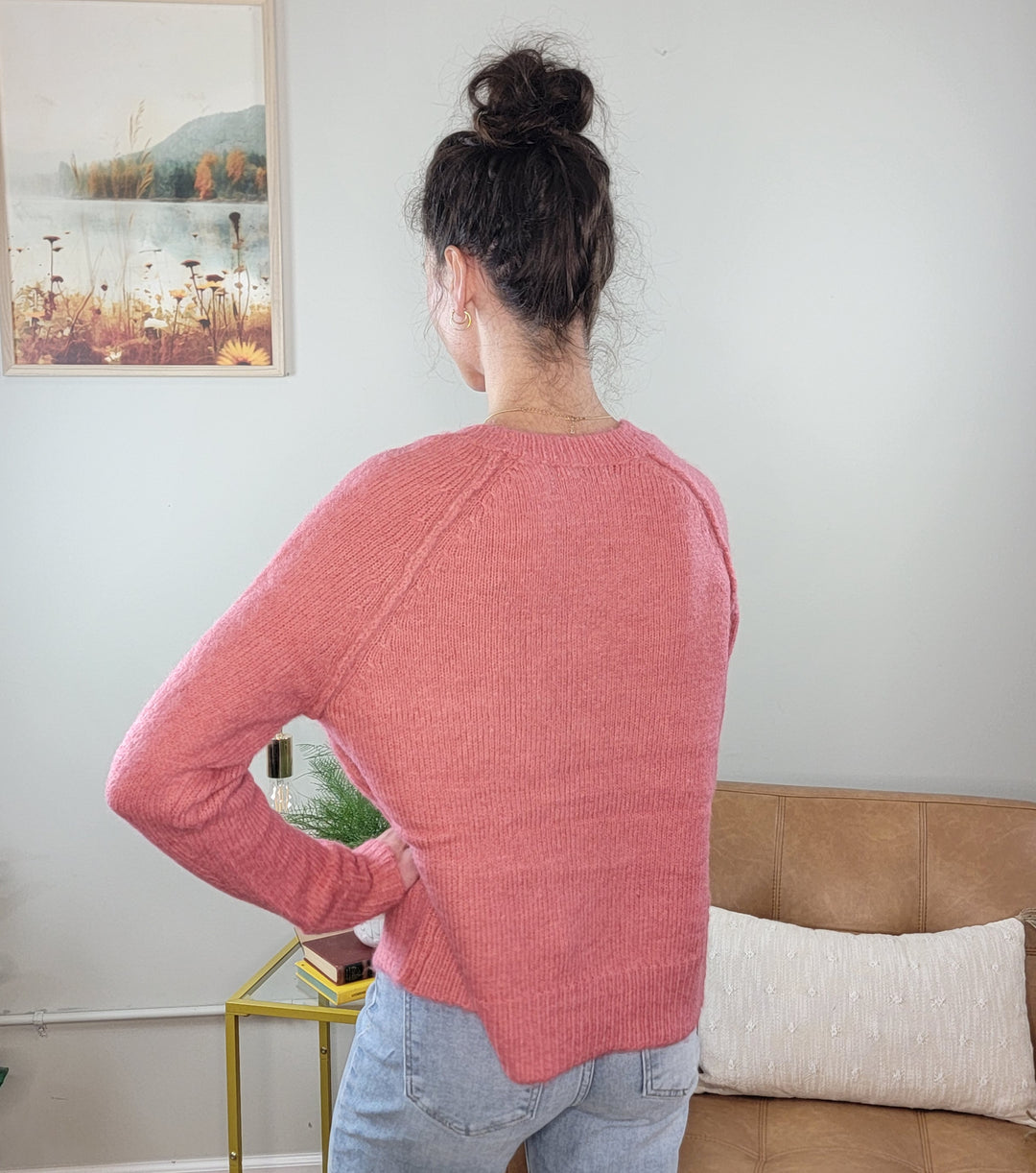 A woman with dark hair in a bun stands with her back to the camera wearing the Staccato Avyana Bow Embellished Knit Sweater in berry and light blue jeans, in a cozy living room with a tan sofa and framed nature picture on the wall.