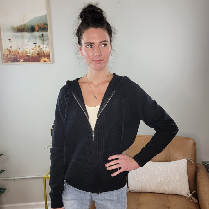 A woman with dark hair in a bun poses indoors, wearing the Staccato Neve Black Knit Zip-Front Hoodie over a light top and light blue jeans. She stands by a tan couch and framed artwork, with one hand on her hip.