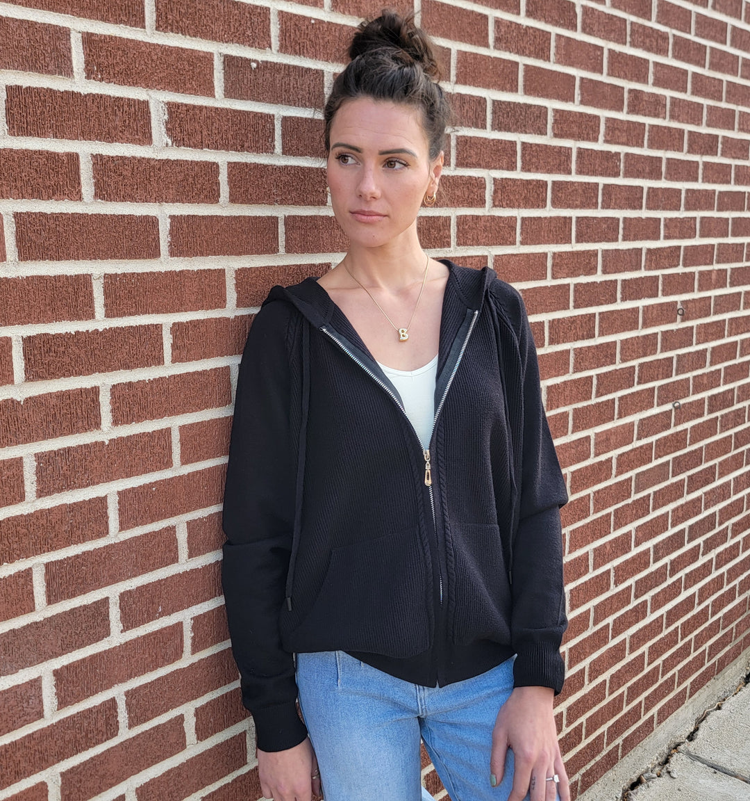 A woman with dark hair styled in a bun stands against a red brick wall, wearing the Staccato Neve Black Knit Zip-Front Hoodie over a white top and light blue jeans, gazing slightly to the side with a neutral expression.