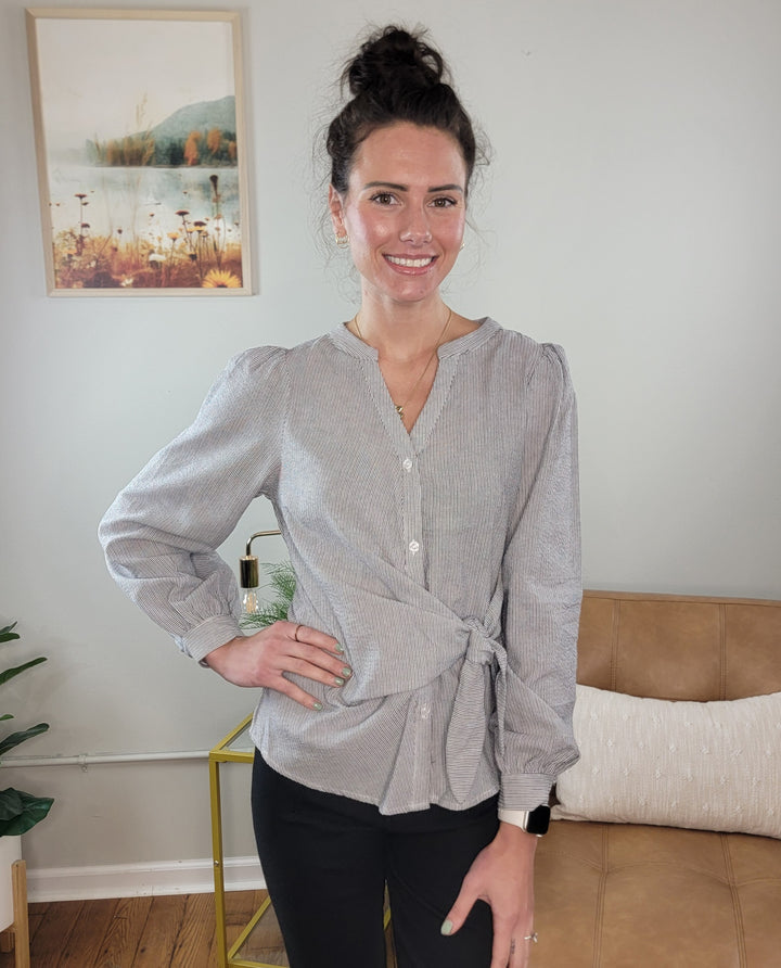 A woman with dark hair in a bun, wearing the Allie Rose Masie Wrap-Front Blouse for Work in light gray and black pants, smiles in a living room with a tan couch, plant, framed art, and yellow side table in the background.