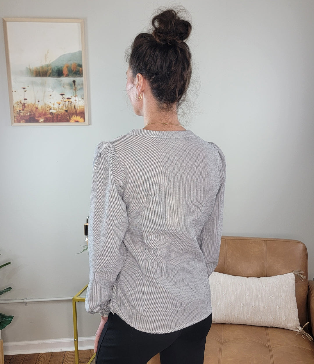 A woman with brown hair in a bun stands indoors facing away, wearing the Allie Rose Masie Wrap-Front Blouse for Work in light gray and black pants, in a modern room with a tan couch, cushion, table, and framed nature wall art.