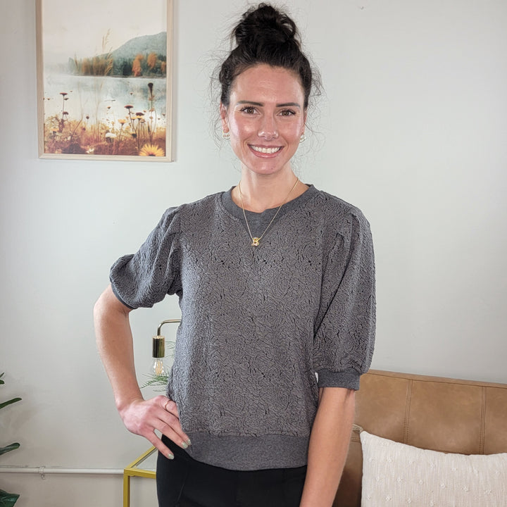 A woman with dark hair in a bun, wearing the Hem &amp; Thread Willa Floral Knit Short Sleeve Top and black pants, stands smiling indoors. Behind her is a framed landscape picture and a tan couch with a white pillow.