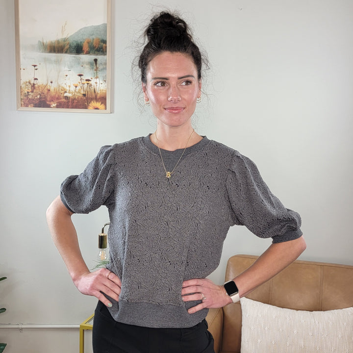 A woman with dark hair in a bun stands indoors, wearing the Hem &amp; Thread Willa Floral Knit Short Sleeve Top and black pants, accessorized with a necklace and smartwatch. She smiles slightly with her hands on her hips; a framed picture hangs behind her.