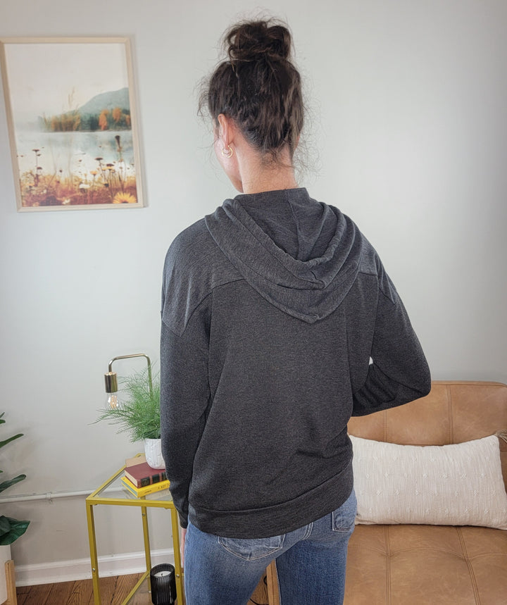 A person with dark hair in a bun wears the Staccato Aviana Fabric Blocked Sweatshirt and blue jeans, standing with their back to the camera in a cozy room featuring a couch, plant, lamp, and wall art.