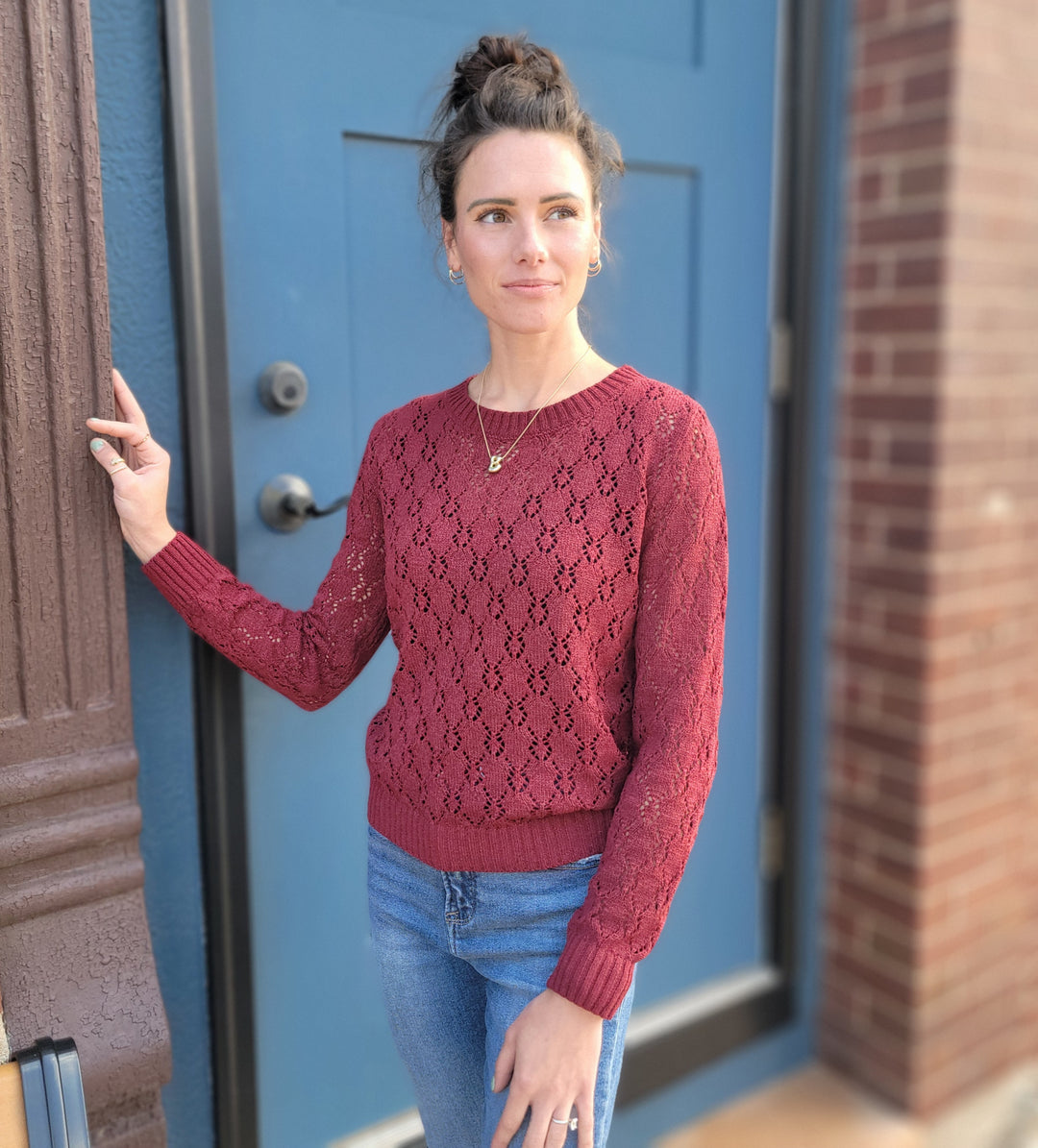 A woman with brown hair in a bun wears the Allie Rose Dixie Pointelle Open Knit Sweater in burgundy and blue jeans, standing by a blue door and brick wall, gazing softly to the side.