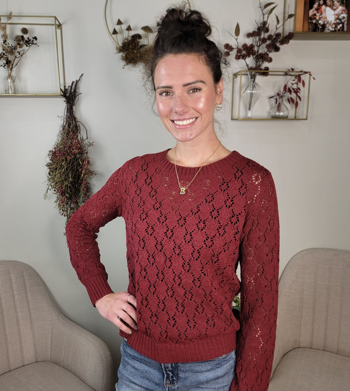 A woman with dark hair in a bun, wearing the Allie Rose Dixie Pointelle Open Knit Sweater and jeans, stands smiling with one hand on her hip in a cozy room decorated with dried flowers and beige chairs.