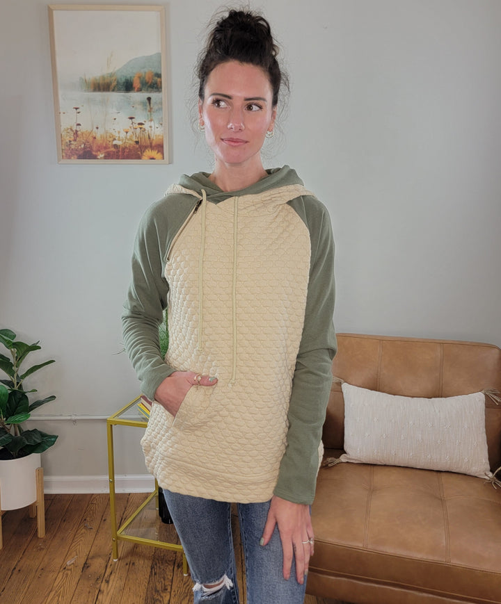A woman with dark hair in a bun wears the Doe &amp; Rae Inaya Quilted Double Hoodie, featuring green sleeves and tan quilted fabric, paired with blue jeans. She stands indoors by a tan couch, with plants and artwork in the background.
