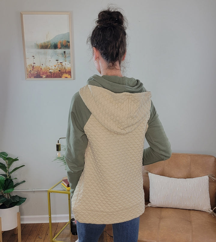 A woman with her hair in a bun stands indoors facing away, wearing the Doe &amp; Rae Inaya Quilted Double Hoodie in green with a textured beige hood. She&