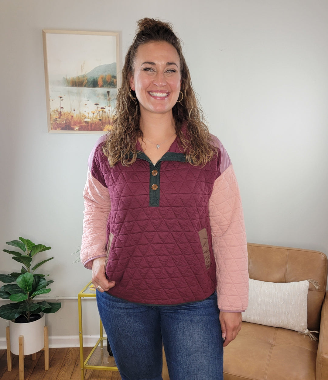 A smiling woman with curly hair stands indoors in the Alaiya Color Blocked Quilted Pullover by Staccato, featuring maroon and pink blocks, paired with blue jeans. A tan sofa, green plant, and framed wall art are in the background.