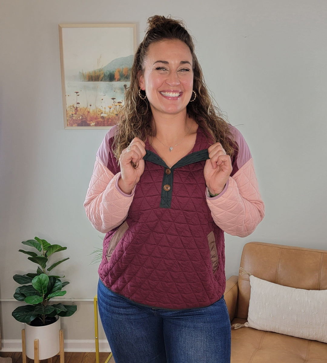 A woman smiles indoors by a tan couch and potted plant, wearing the Staccato Alaiya Color Blocked Quilted Pullover in maroon and pink with blue jeans. A framed nature photo hangs on the wall behind her.