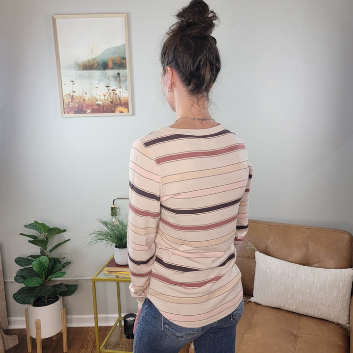 A woman with brown hair in a messy bun, wearing the Thread & Supply Stacy Top and jeans, stands facing away in a cozy room with plants, a tan couch, and landscape wall art.