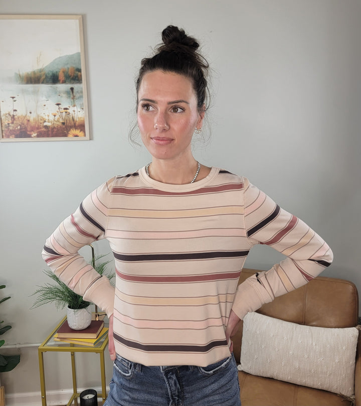 A woman with dark hair in a bun stands indoors, smiling softly with her hands on her hips. She wears the Stacy Top by Thread & Supply with fall stripes and jeans, against a backdrop of wall art and indoor plants.