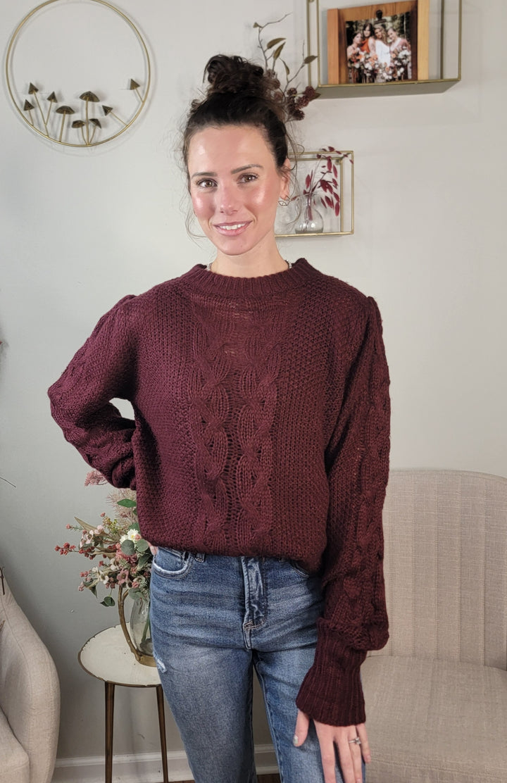 A woman with brunette hair in a bun wears the Doe &amp; Rae Peyton Sweater, a dark burgundy cable knit piece, and blue jeans while standing indoors by a light chair, small table with flowers, and wall decor. She smiles at the camera.