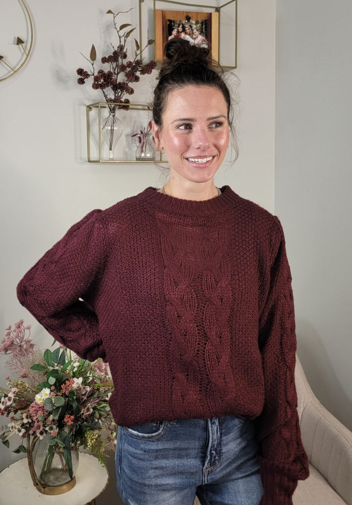 A woman with brown hair in a top bun smiles while wearing the Doe &amp; Rae Peyton Sweater in maroon and blue jeans. She stands indoors beside a table with flowers and a wall shelf featuring decorative items behind her.