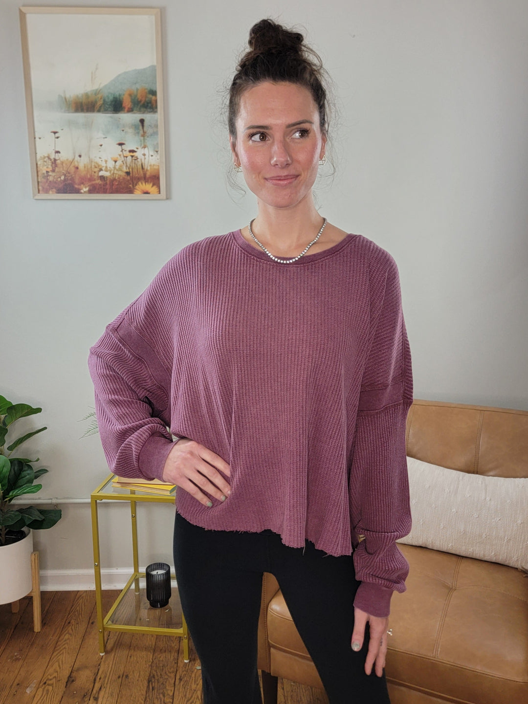 A woman stands indoors smiling softly, wearing the HYFVE, Inc Alma Knit Long Sleeve Top in mauve with black leggings. Her hair is in a bun, and she accessorizes with a necklace. Behind her are a couch, plant, and framed landscape picture.