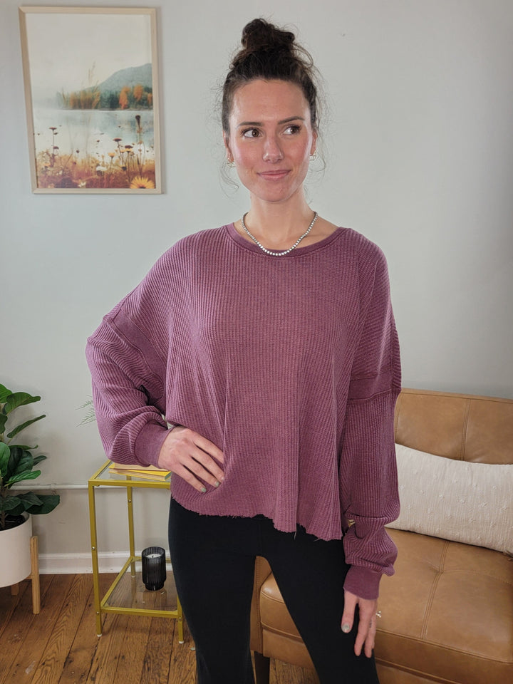 A woman stands indoors smiling softly, wearing the HYFVE, Inc Alma Knit Long Sleeve Top in mauve with black leggings. Her hair is in a bun, and she accessorizes with a necklace. Behind her are a couch, plant, and framed landscape picture.