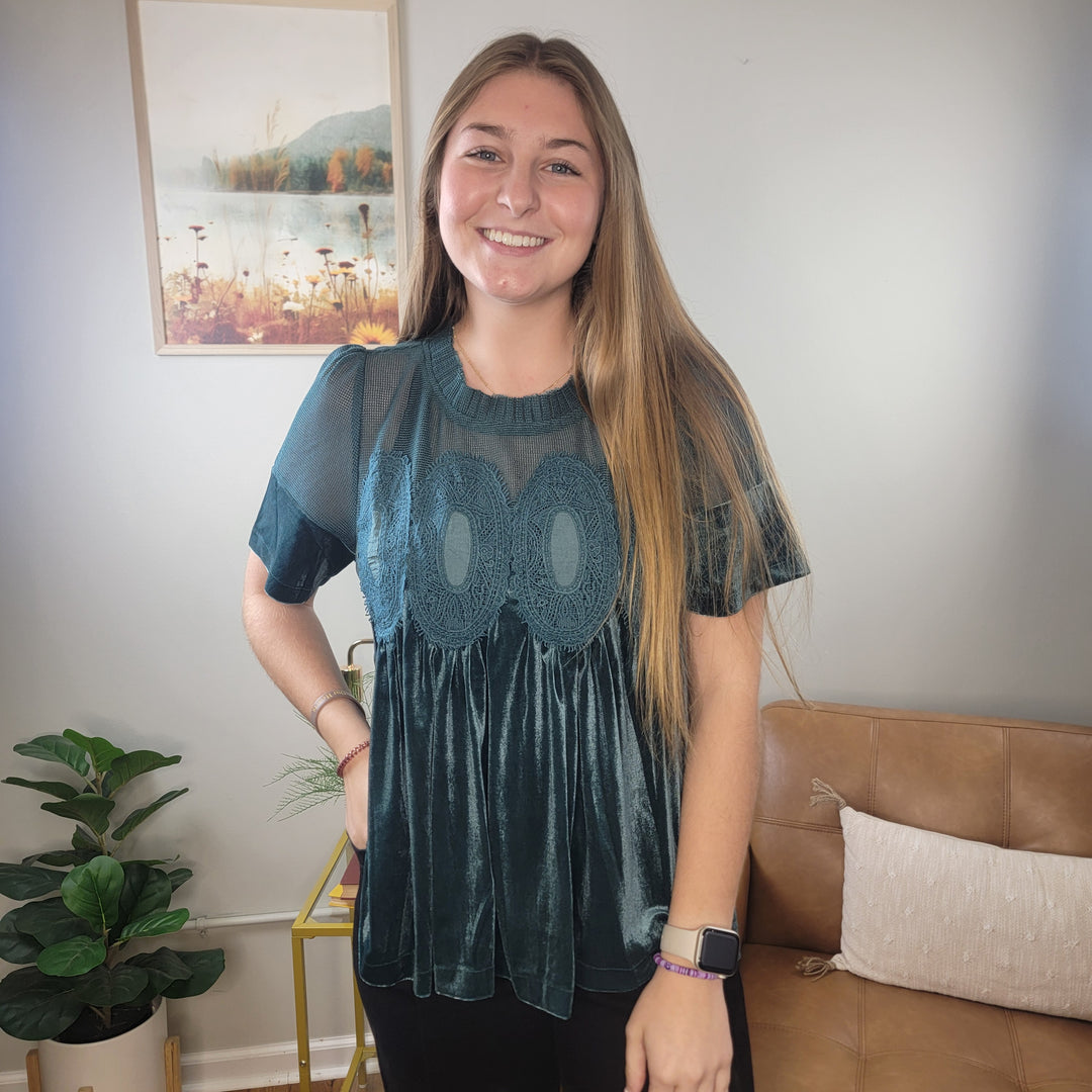 A young woman with long hair stands smiling in a living room, wearing the Fate Preslee Velvet and Lace Babydoll Top. Behind her are a tan couch, a green potted plant, and a framed landscape painting on the wall.