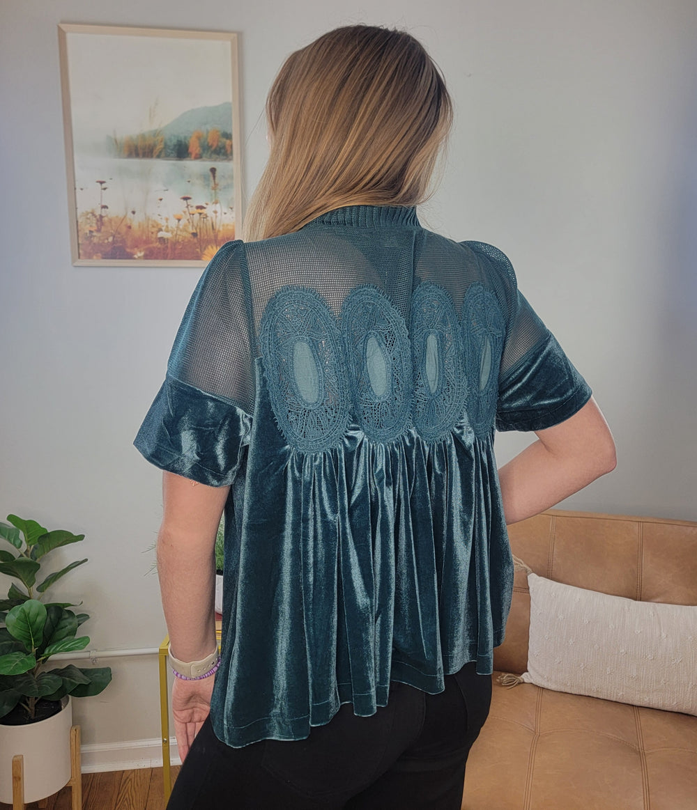 A woman with long, light brown hair stands indoors with her back to the camera, wearing the Fate Preslee Velvet and Lace Babydoll Top in teal. The room features a plant, tan sofa, and framed picture on the wall.
