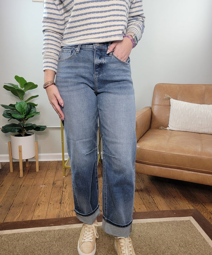 A person wearing Risen Mira Cropped Barrel Jeans, a striped sweater, and beige sneakers stands on a rug in a living room with a tan sofa, pillow, and potted plant. Their head is out of the frame.